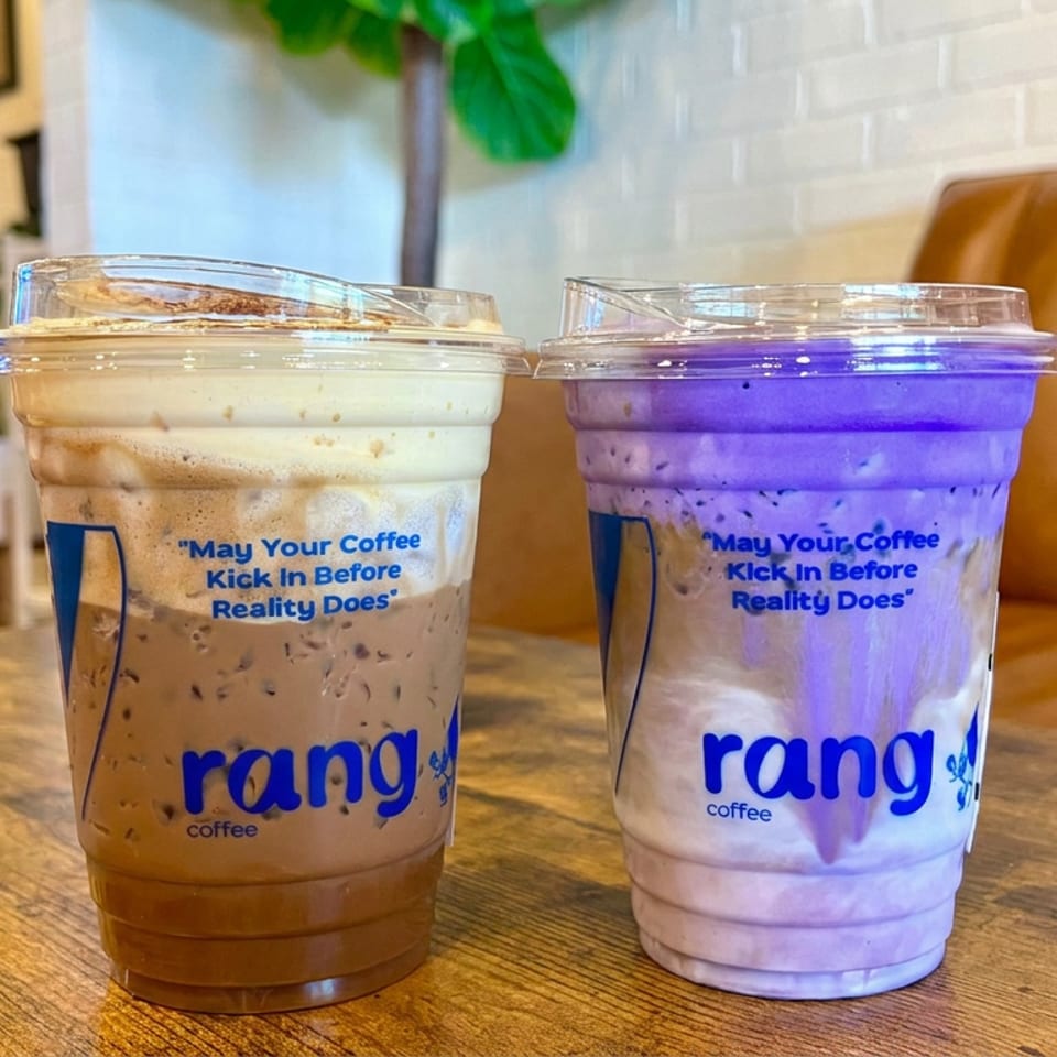 Two iced drinks in branded Rang Coffee cups sit on a wooden table, with a cozy café booth and plants behind them.