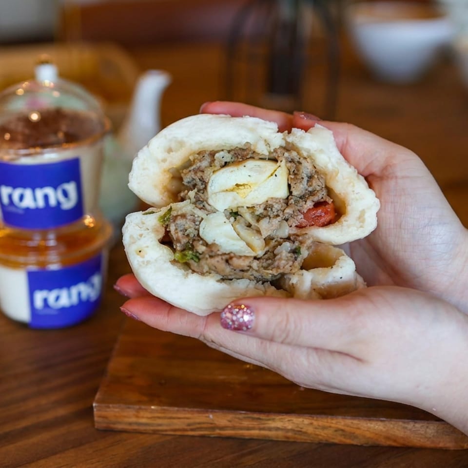 Hands hold a stuffed steamed bun filled with meat, egg, vegetables, and sauce beside branded tea cups on a wooden table.